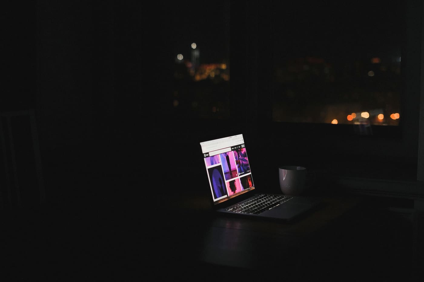 A laptop glowing on a desk in a dark room at night with city lights outside the window